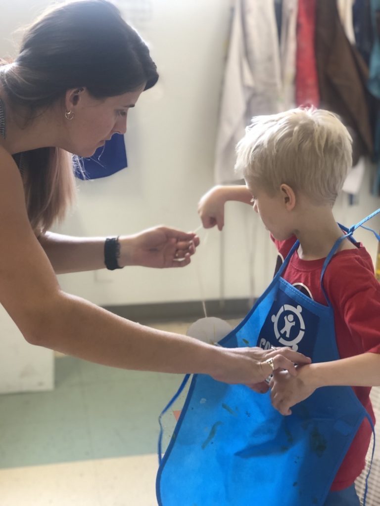 A volunteer helping a child put on a Connors Heroes apron for an art session