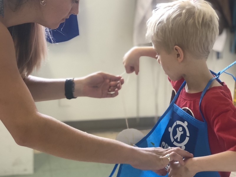 A volunteer helping a child put on a Connors Heroes apron for an art session