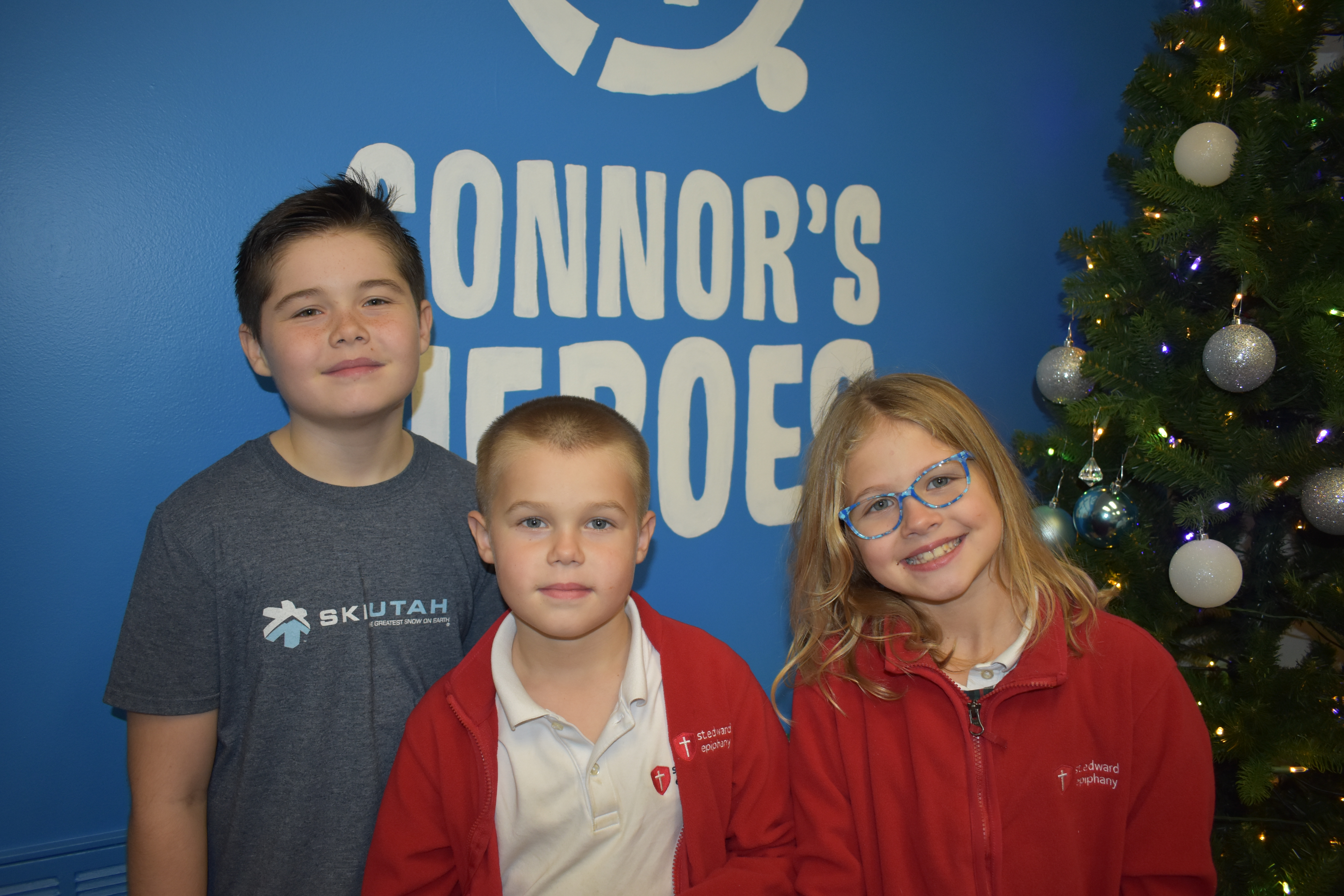 Three siblings smiling and standing in front of the Connor's Heroes logo wall