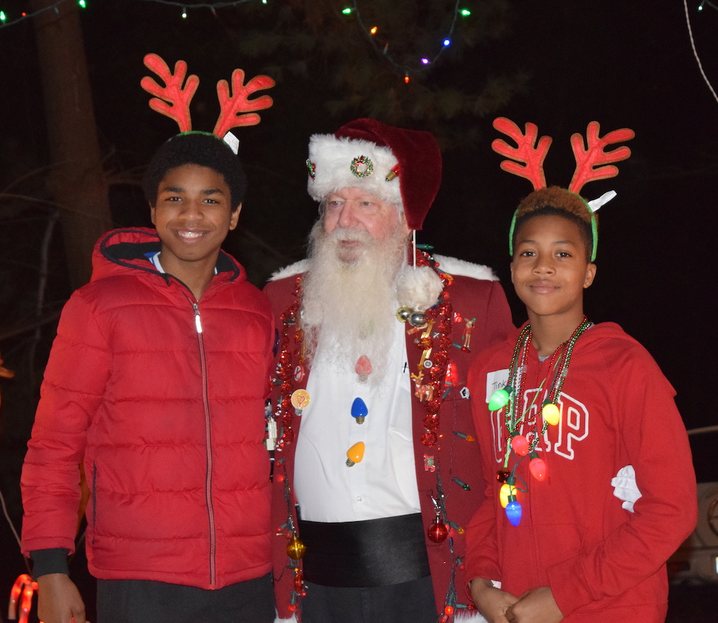 Two teens standing with Santa in front of a house decorated with colorful holiday lights