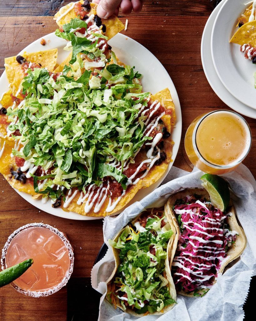 Overhead photo of two plates of nachos and tacos with drinks.