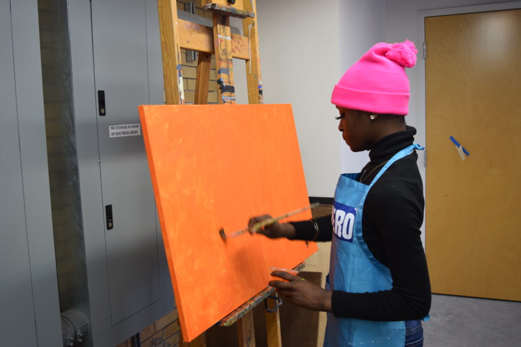 Girl wearing a pink hat painting an orange background on a canvas