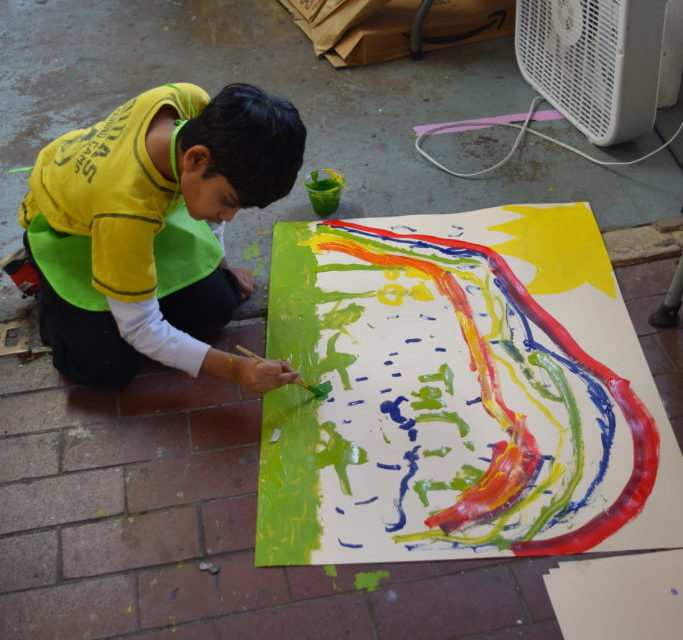 A boy is sitting on the floor painting a rainbow on a large piece of paper