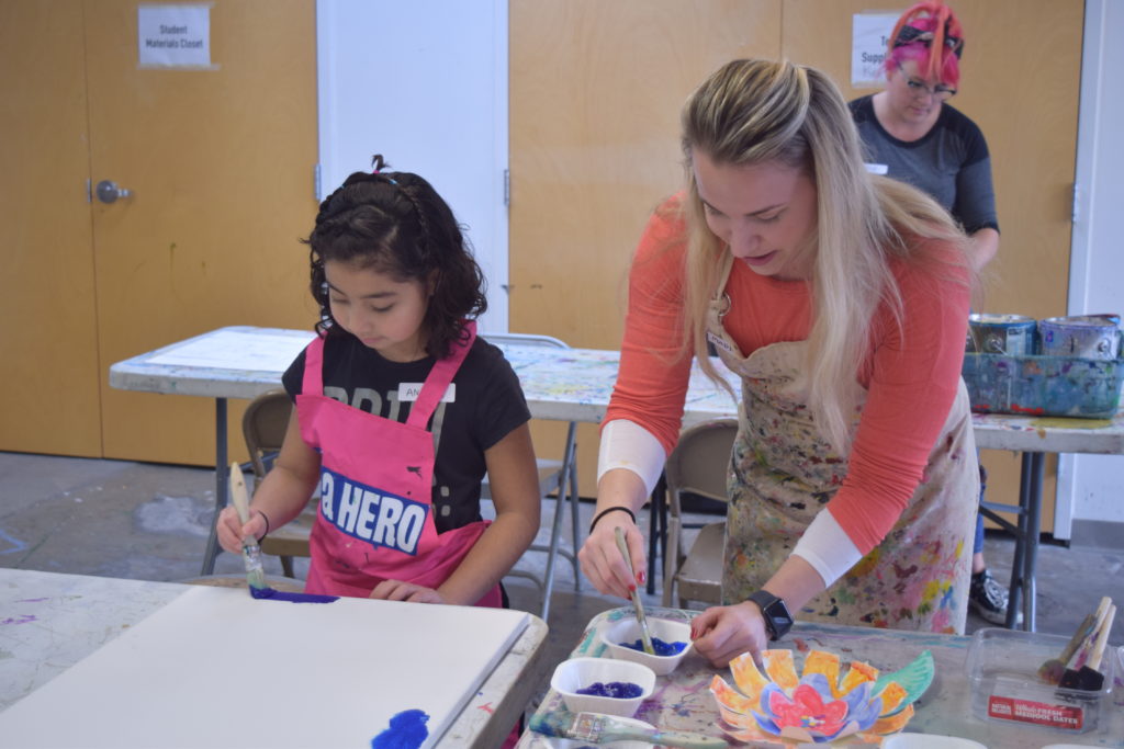 An artist helps a girl paint blue on a large canvas at the Connors Heroes Art Sessions