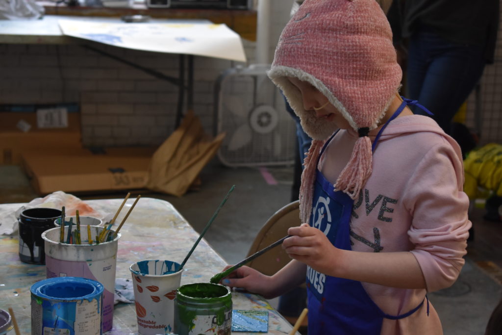 A girl wearing a hat uses green paint to paint a tile at the Connors Heroes Art Sessions