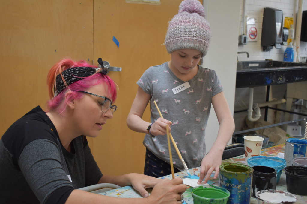 An artist with pink hair helps a girl wearing a hat paint tiles