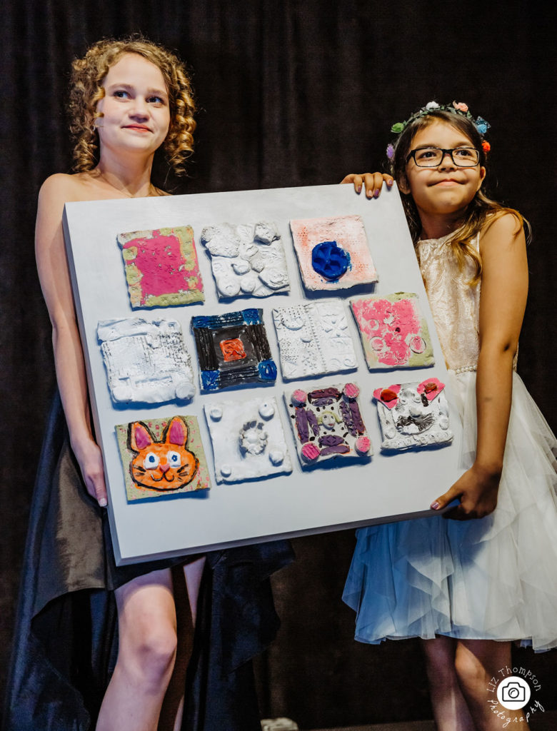 Two girls standing on a stage holding a collage made with painted tiles