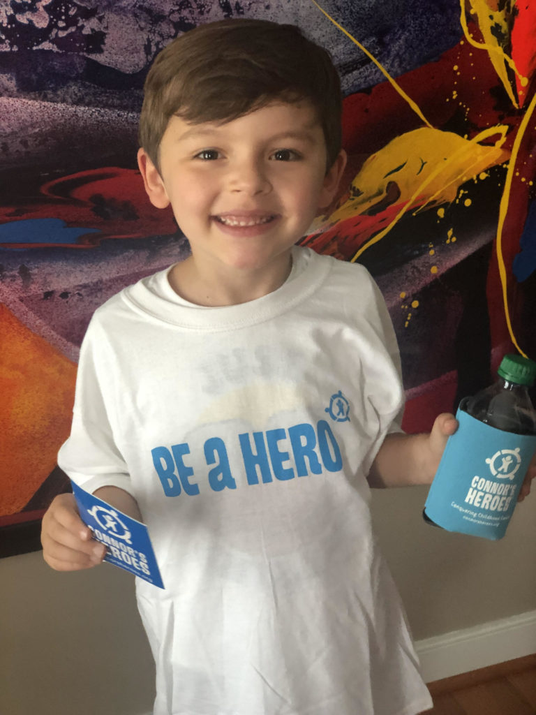 A boy wearing a white Be A Hero t-shirt, holding Connor's Heroes sticker and koozie