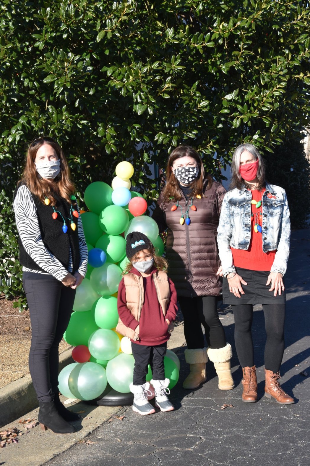 3 women and a child wearing masks standing outside beside a holiday tree made out of green and white balloons
