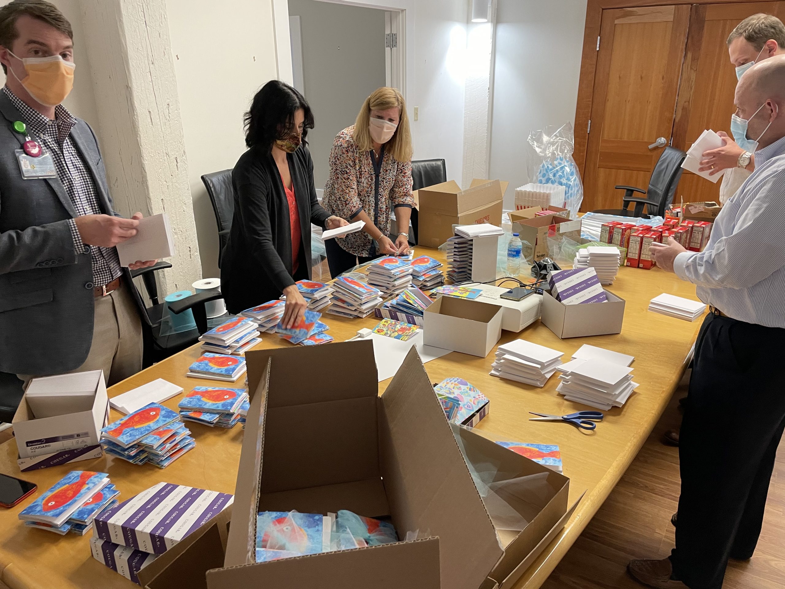 Image 5 people standing around a conference table. On the table are piles of greeting cards and boxes.