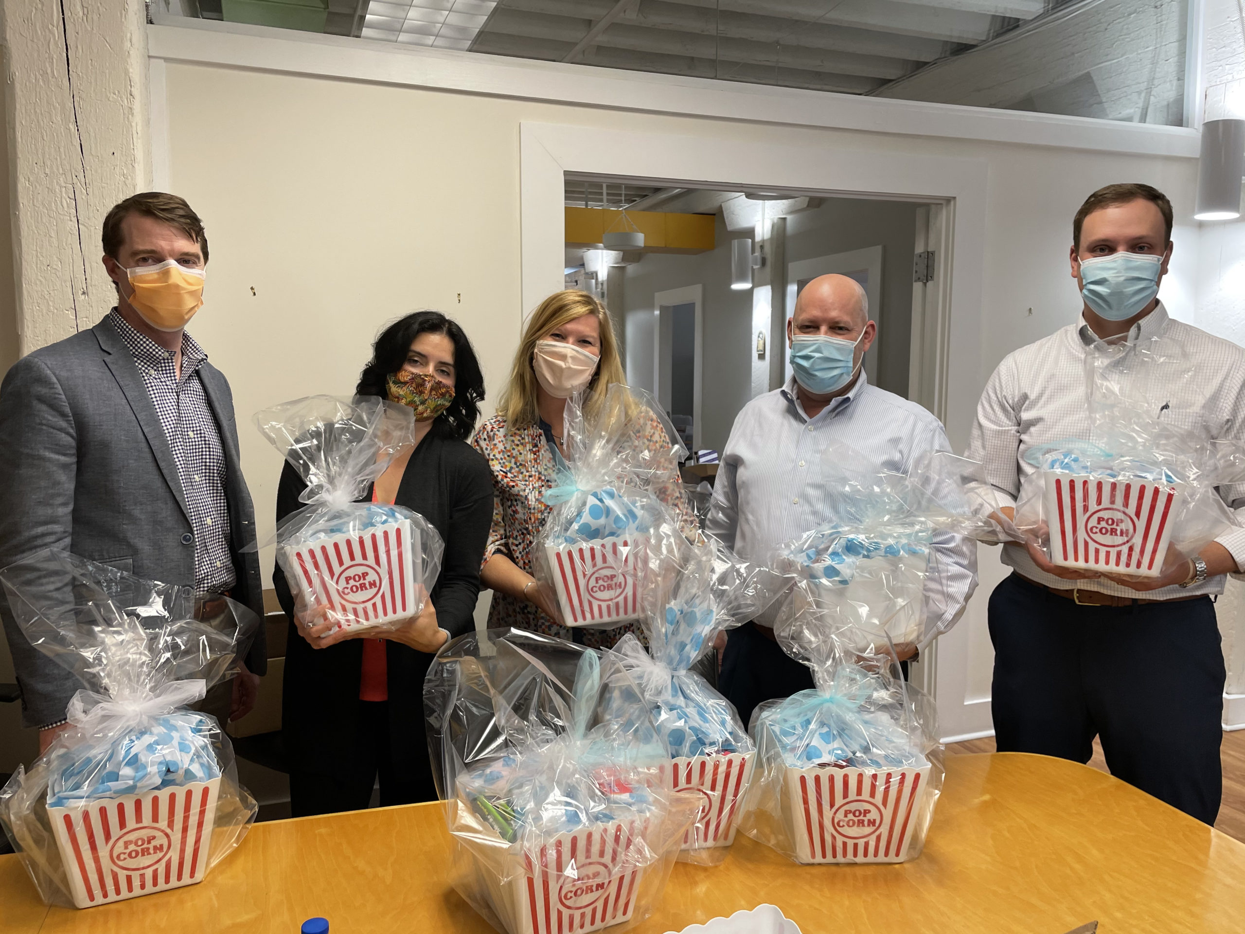 Image 5 people standing behind a large table. On the table are gift baskets wrapped with tissue paper.