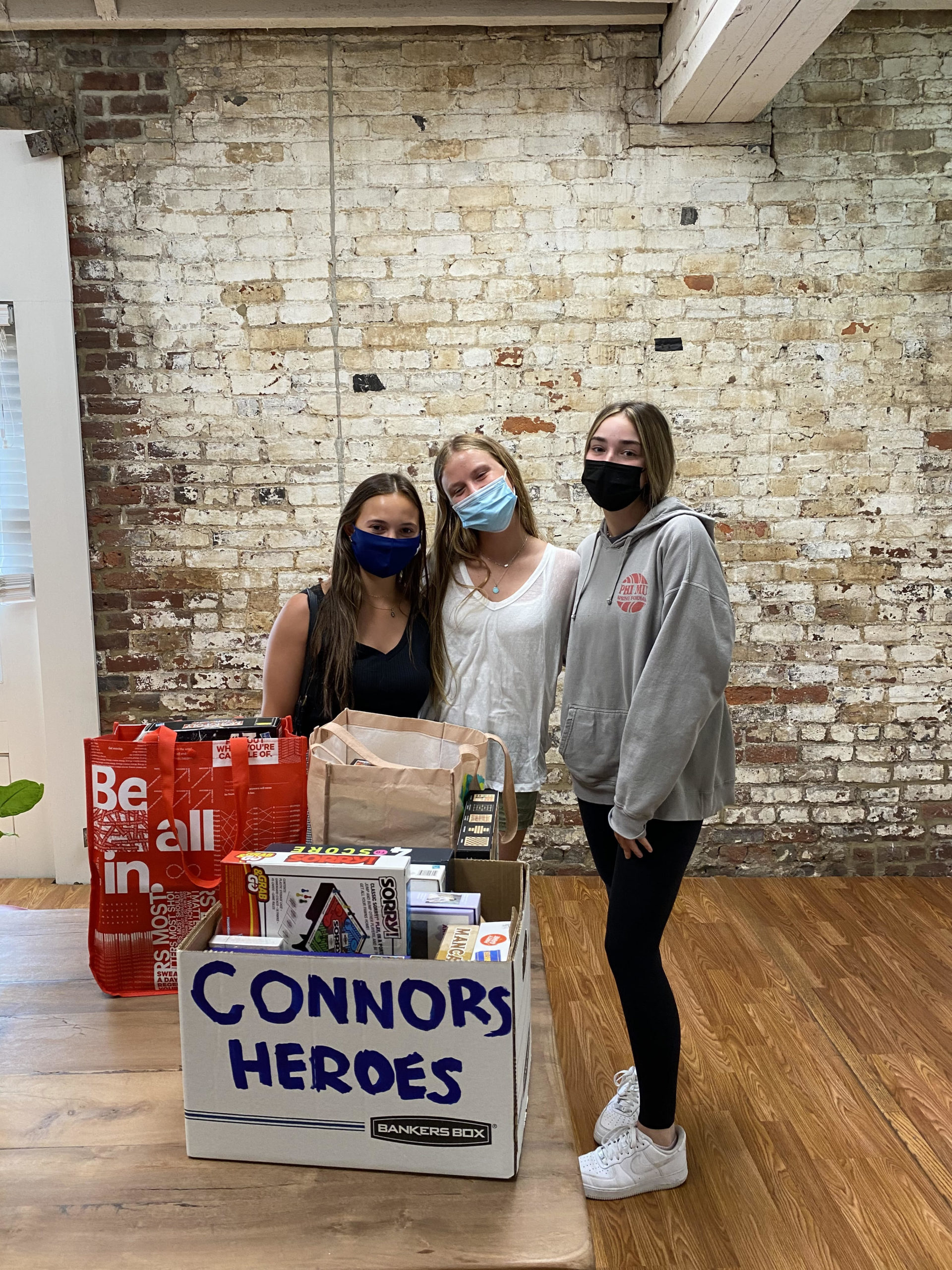 Image three teenagers wearing masks standing behind a table with a box full of board games