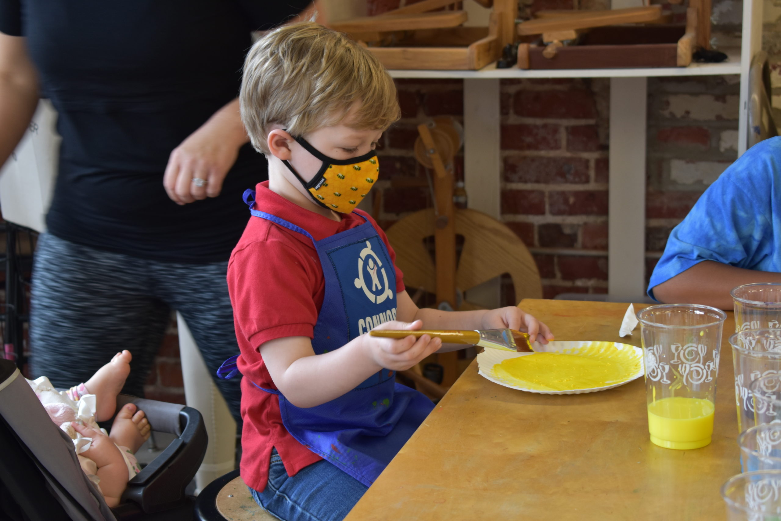 A child wearing a mask sitting at a table painting a paper plate yellow