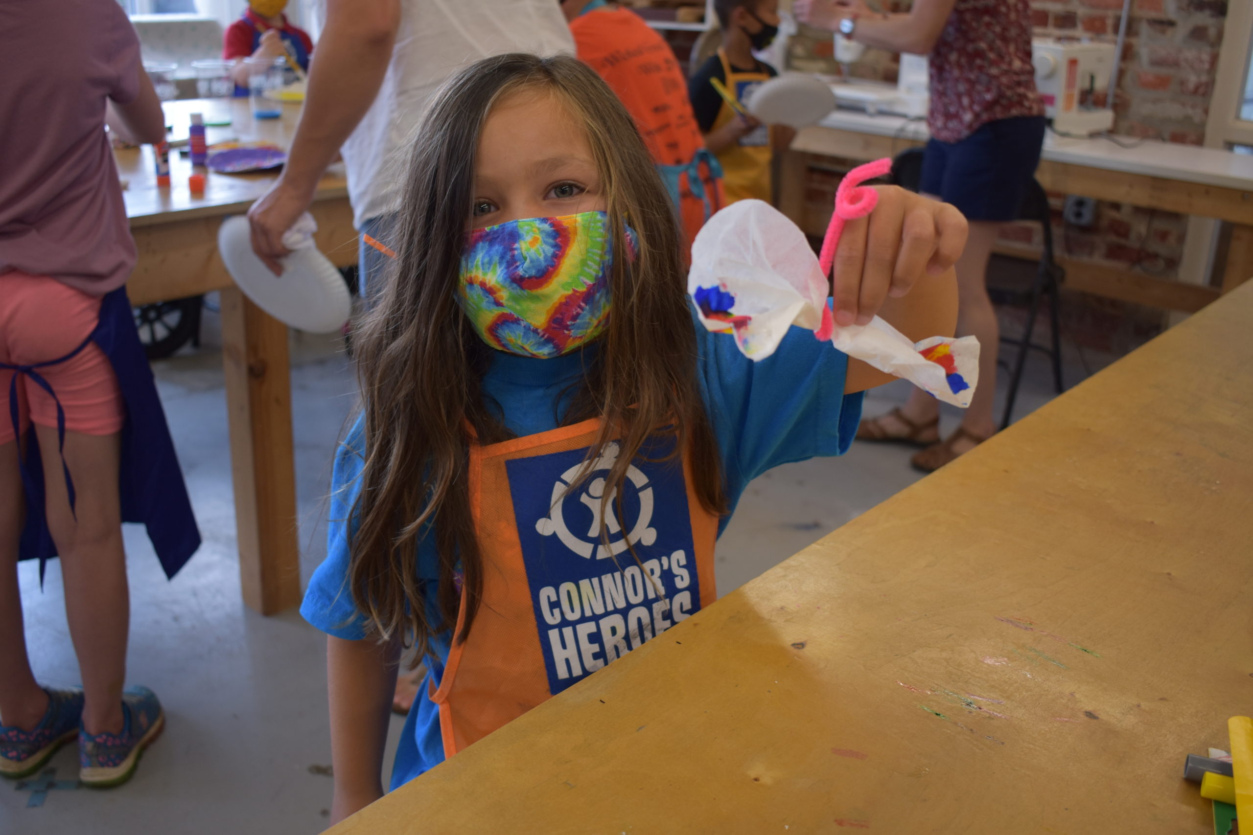 A child wearing a mask holding up a butterfly made out of paper and pipe cleaners