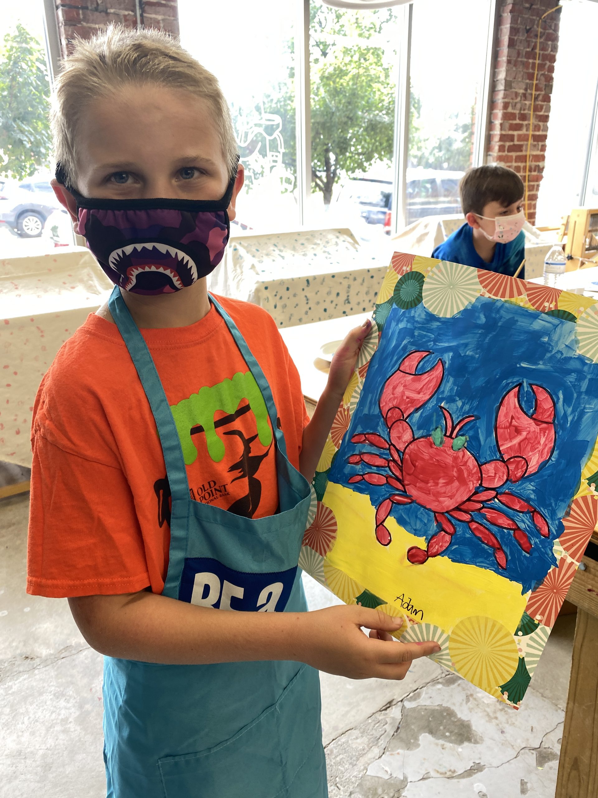 A child wearing a mask and holding up a painting of a crab