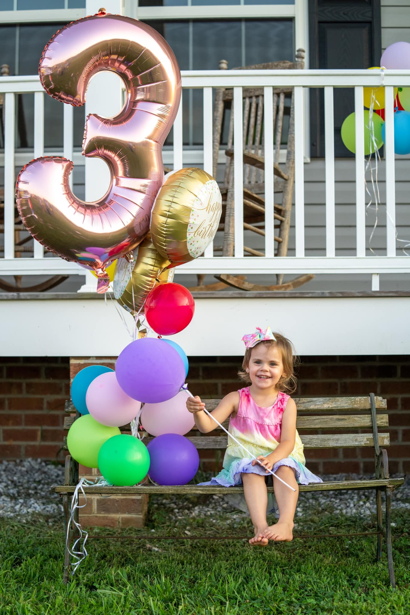A girl sits on a park bench holding a group of balloons one balloon is a gold number three