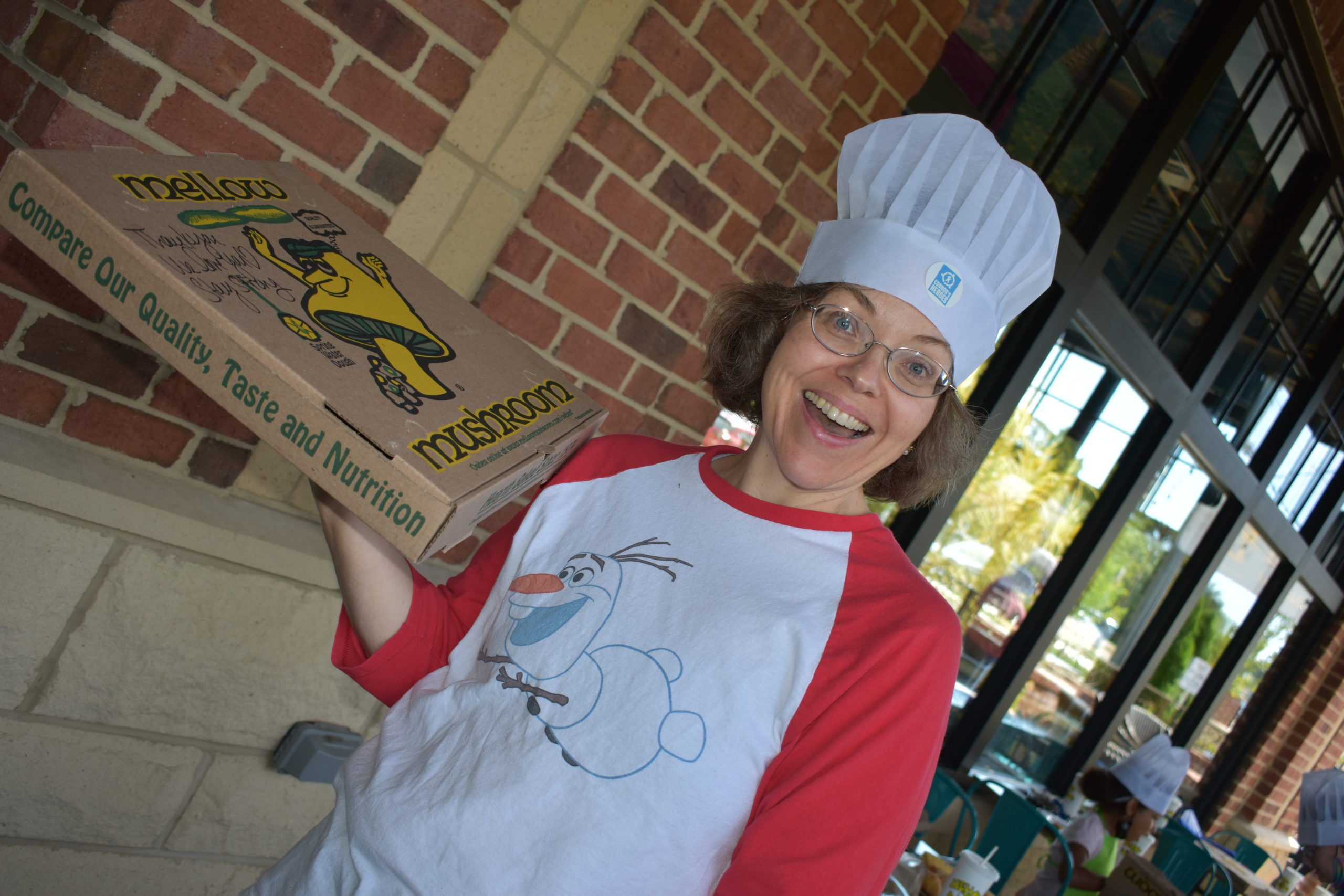 Image a woman wearing a chef hat holding a pizza box and smiling