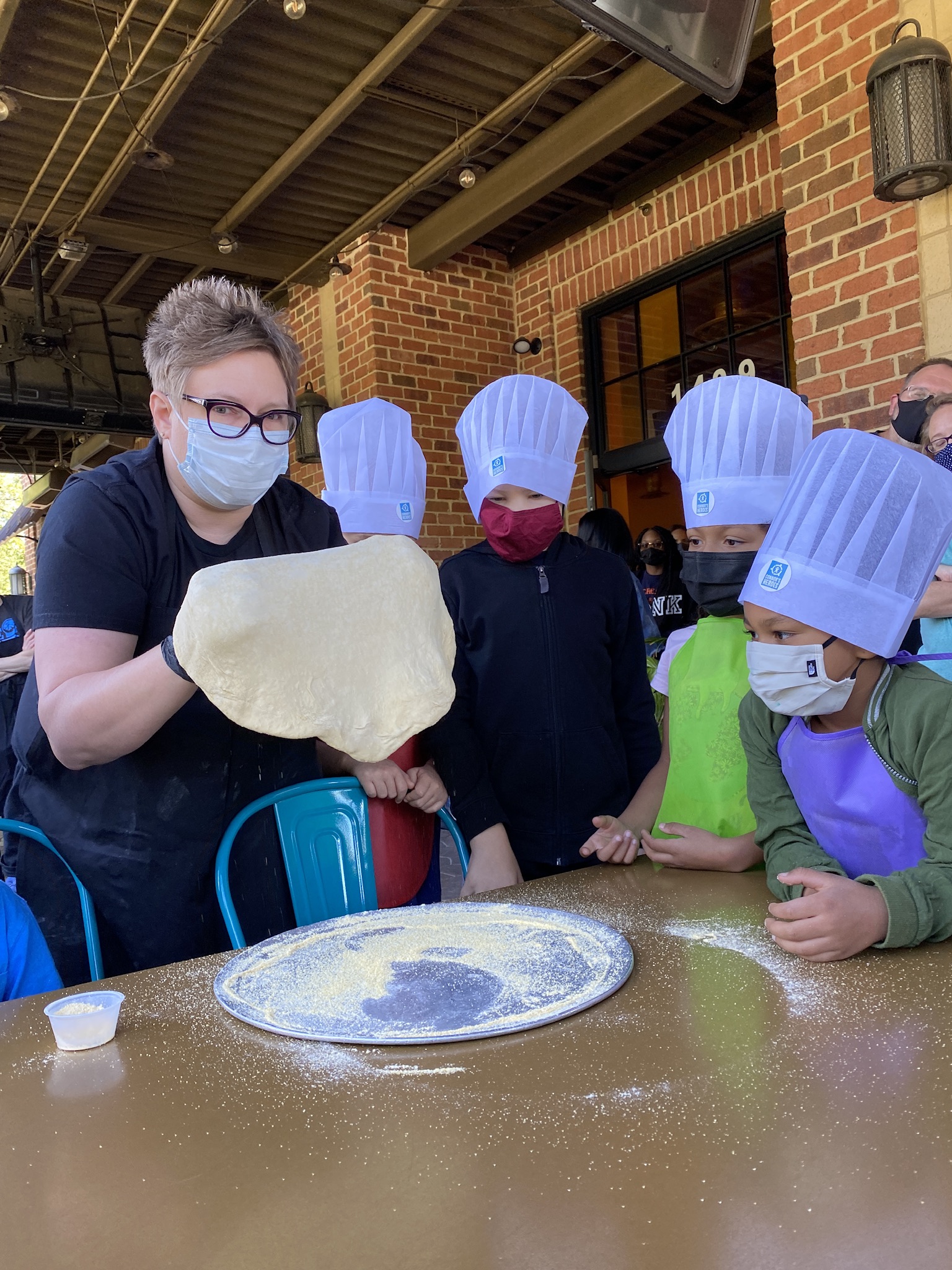Image an adult helping a group of four kids roll a dough of pizza