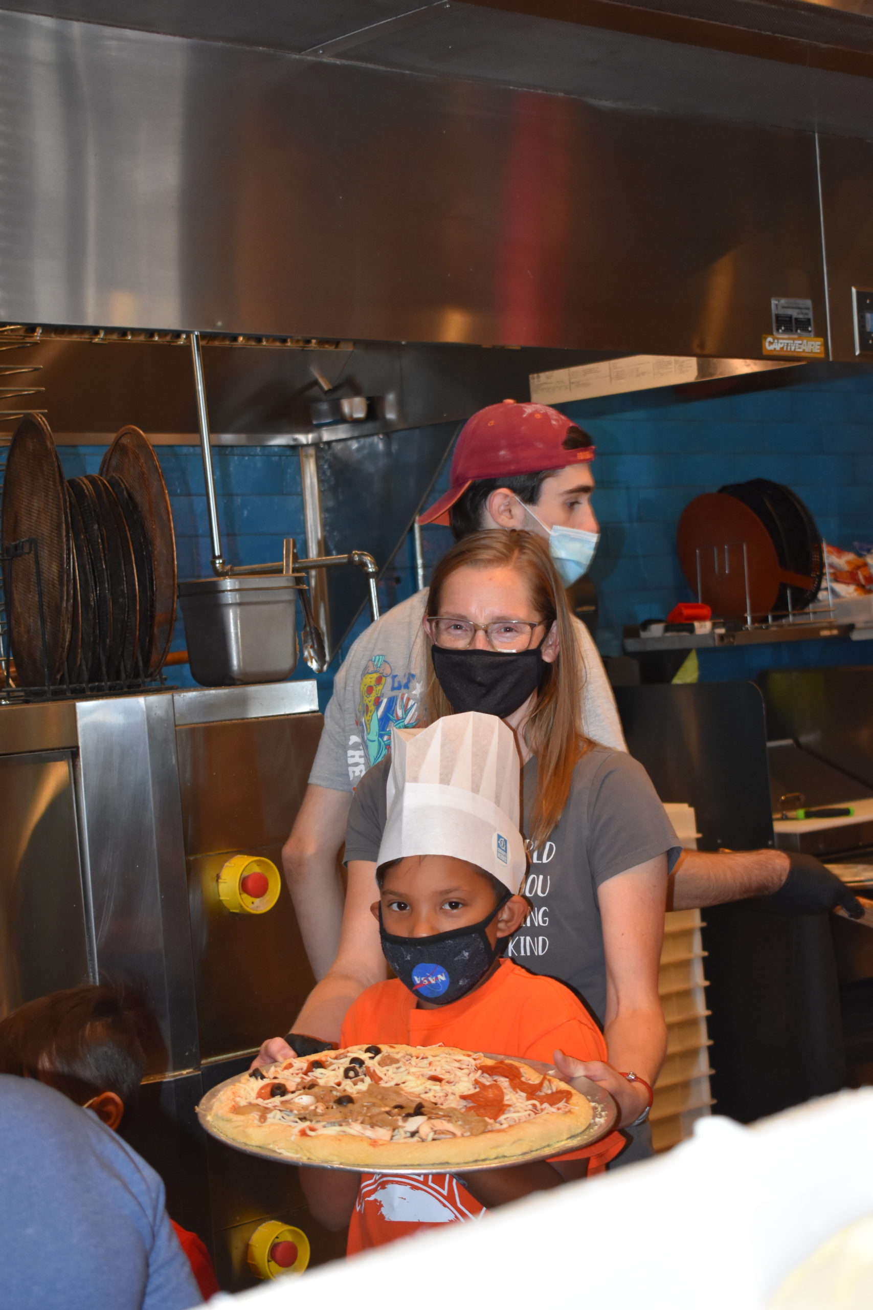 Image a child carrying an uncooked pizza into the kitchen of a restaurant