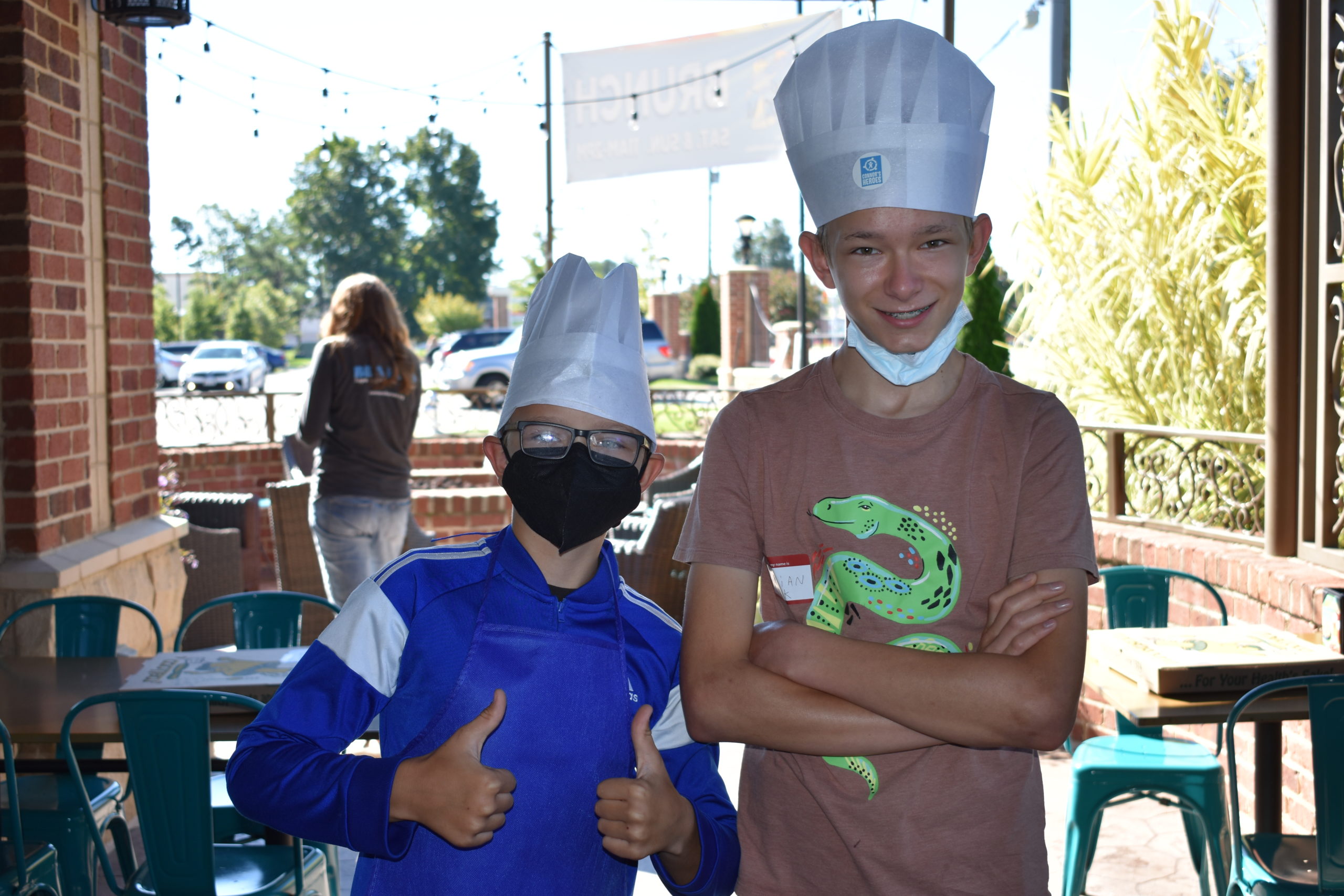 Image two boys wearing chef hats and standing outside at a restaurant