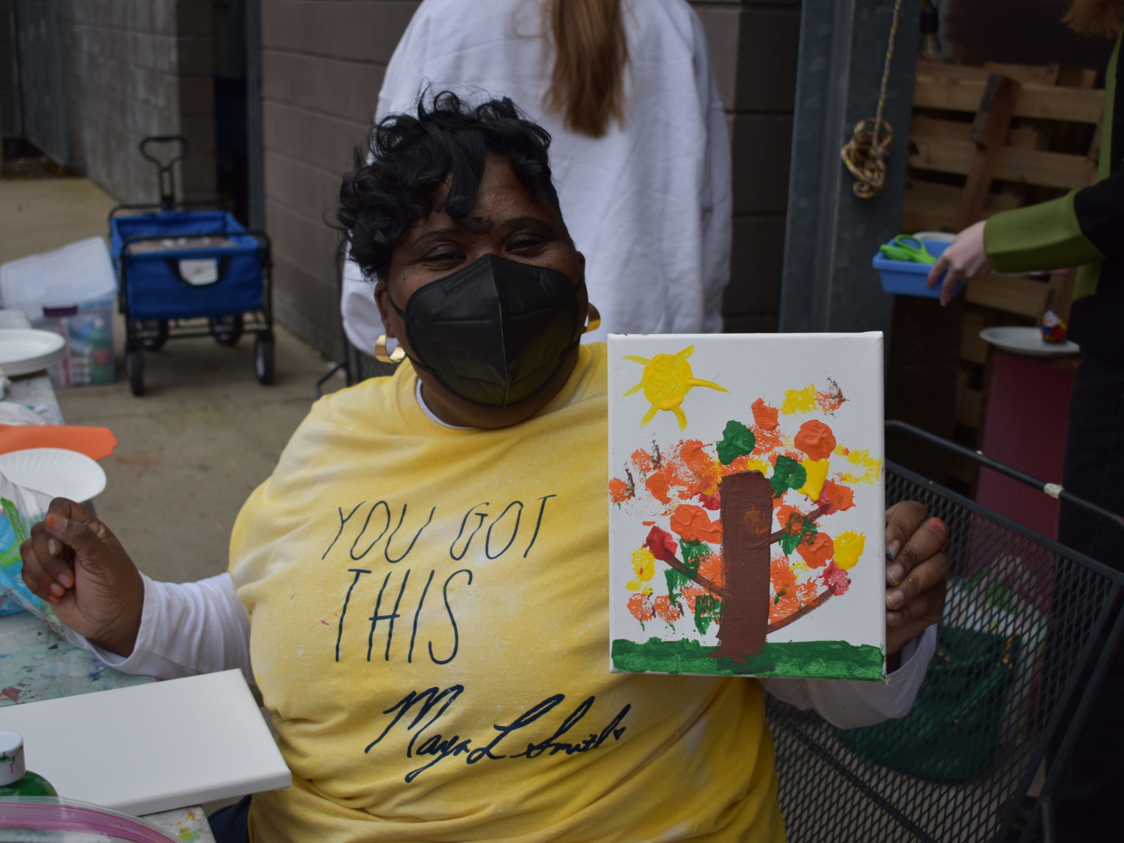 A woman wearing a mask holding up a painting of a tree