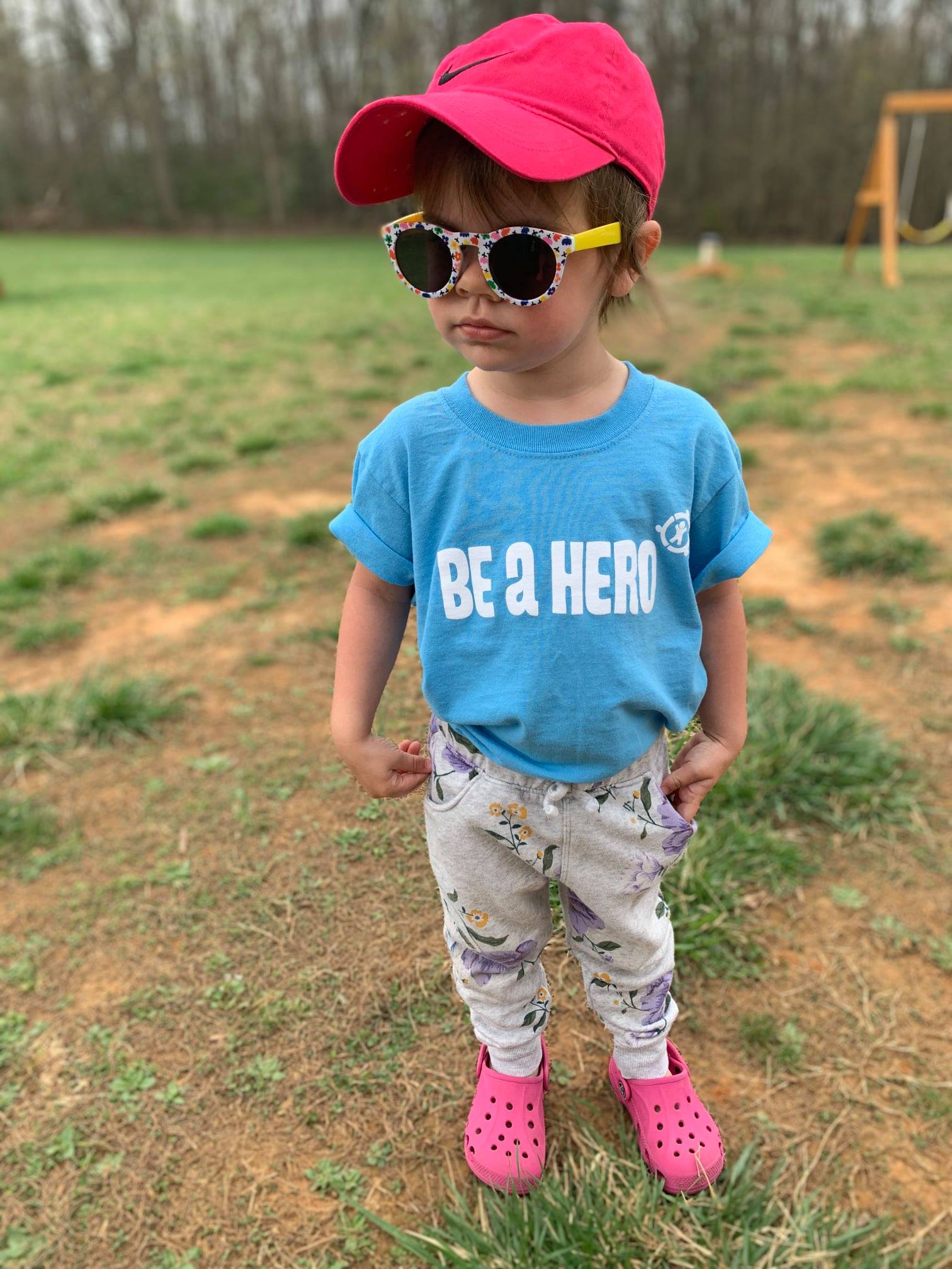 A child wearing a pink hat with matching pink sandals and colorful sun glasses with a bright blue tee shirt that reads Be A Hero