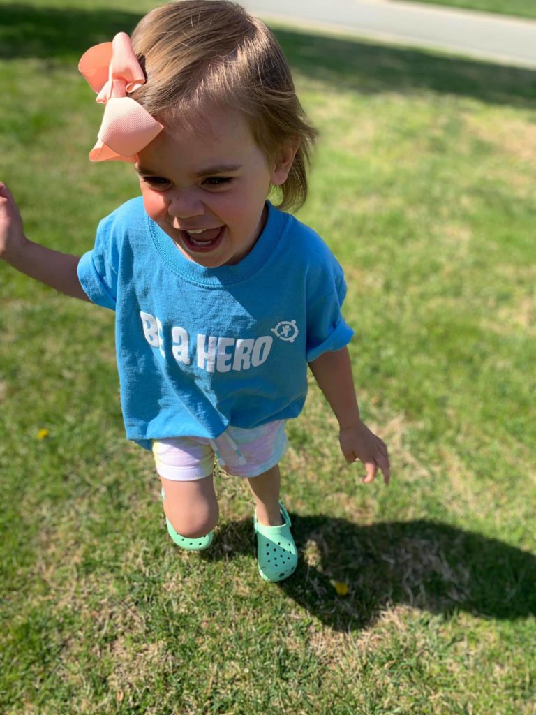 A child with a pink bow in her hair running in a yard wearing a bright blue tee shirt that reads Be A Hero