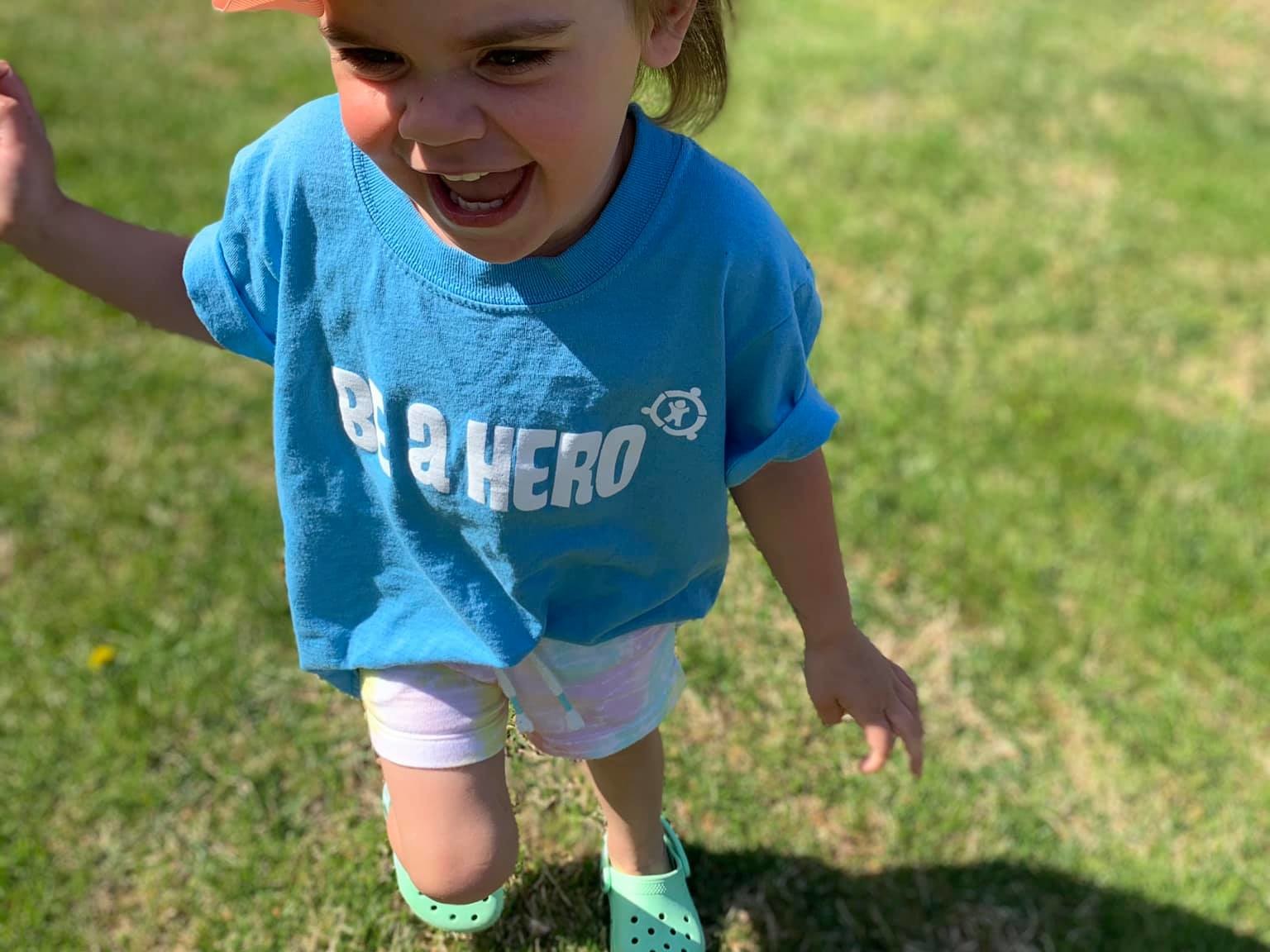 A child with a pink bow in her hair running in a yard wearing a bright blue tee shirt that reads Be A Hero