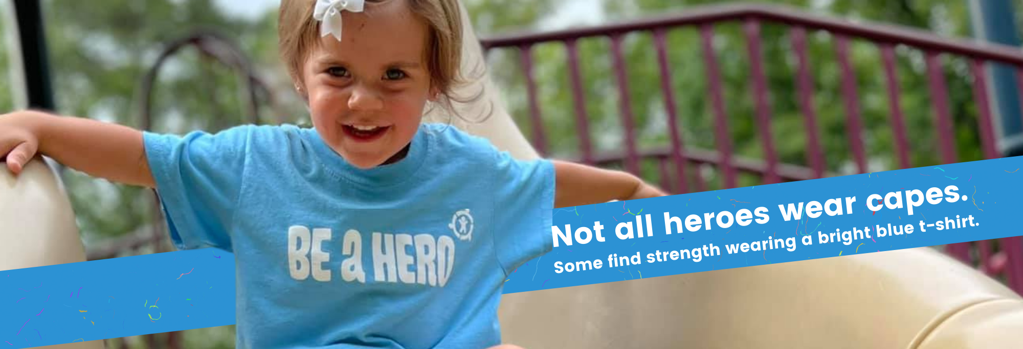 A child with a pink bow in her hair going down a slide wearing a bright blue tee shirt that reads Be A Hero The text reads Not all heroes wear capes. Some find strength wearing a bright blue t-shirt.