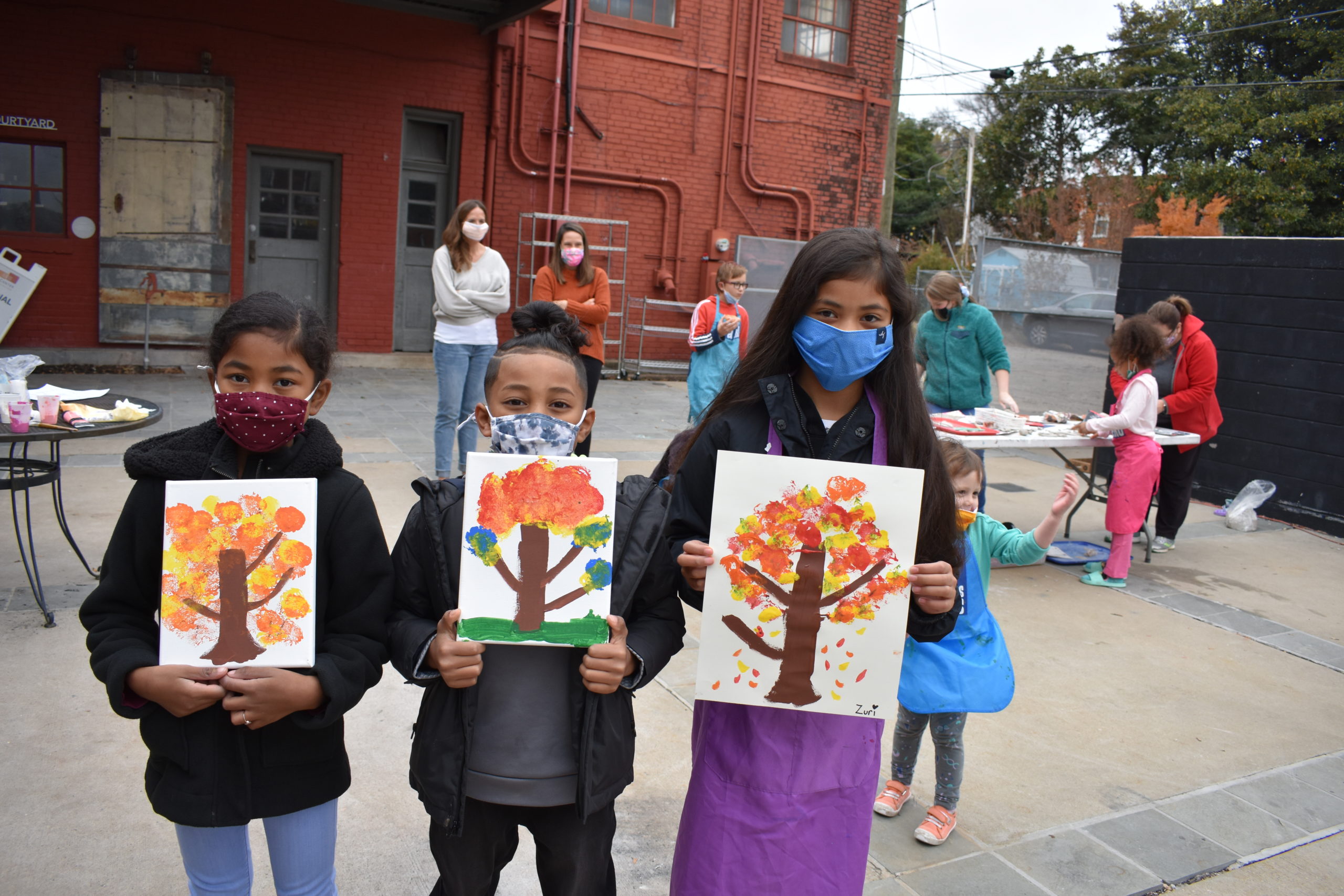 Three children standing in a row holding up paintings of trees