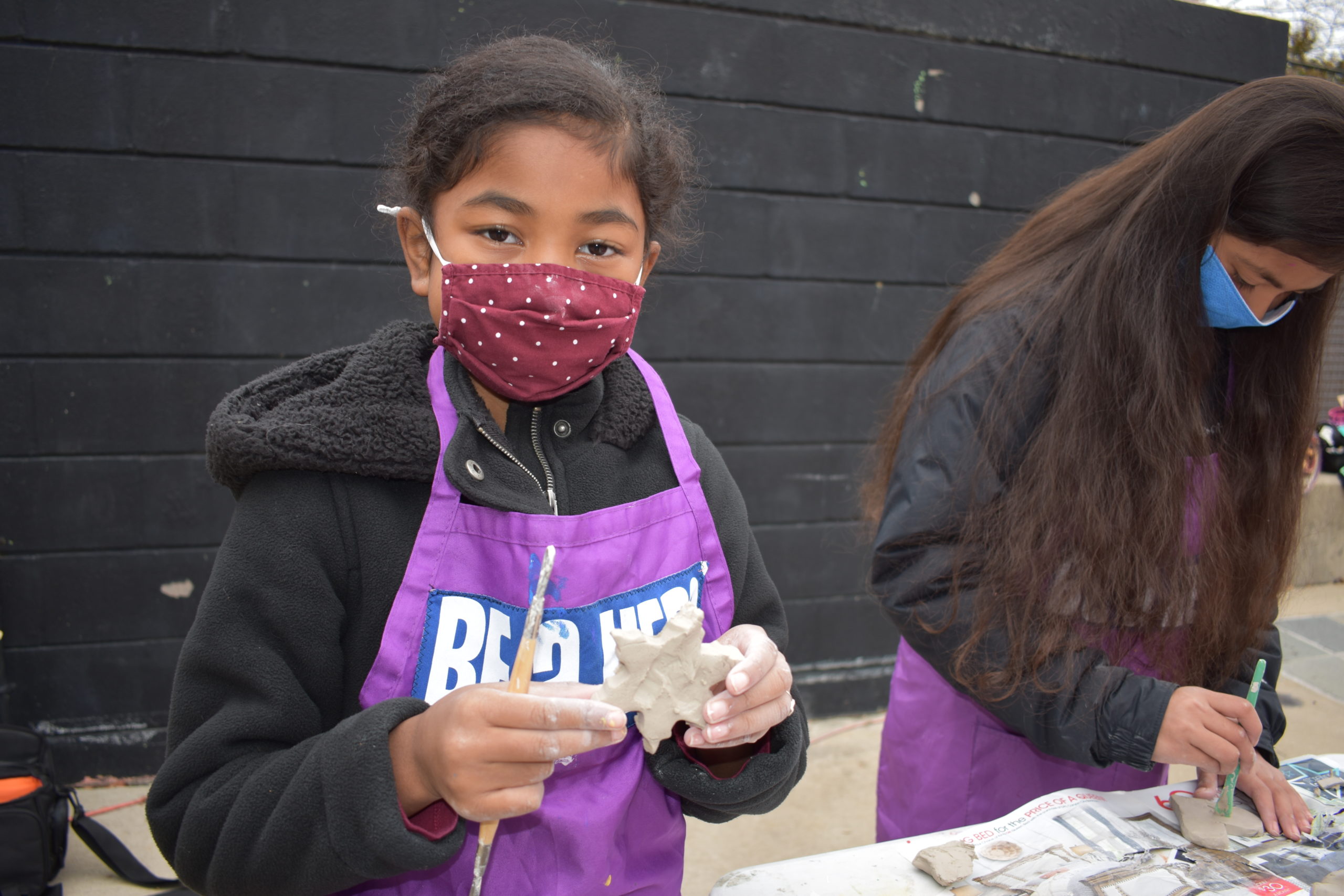 A girl wearing a cloth face mask looking at the camera holding a snowflake made out of clay