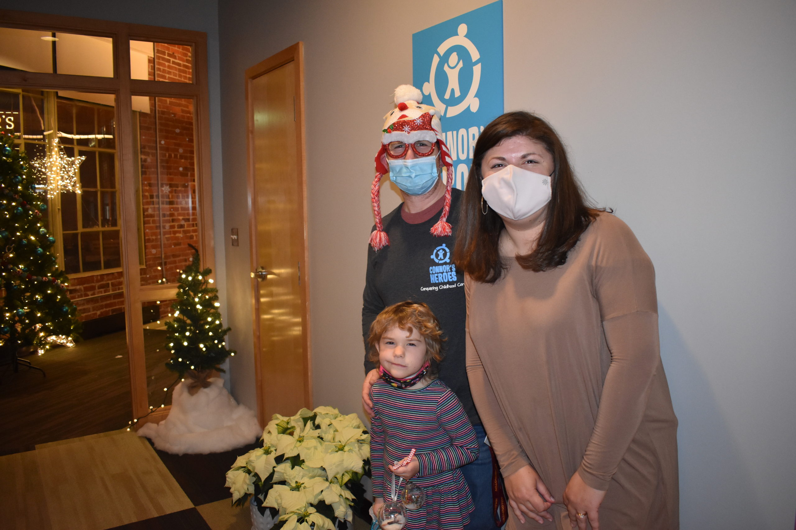A man wearing a holiday hat is standing with a woman and a girl in an office decorated for a holiday party