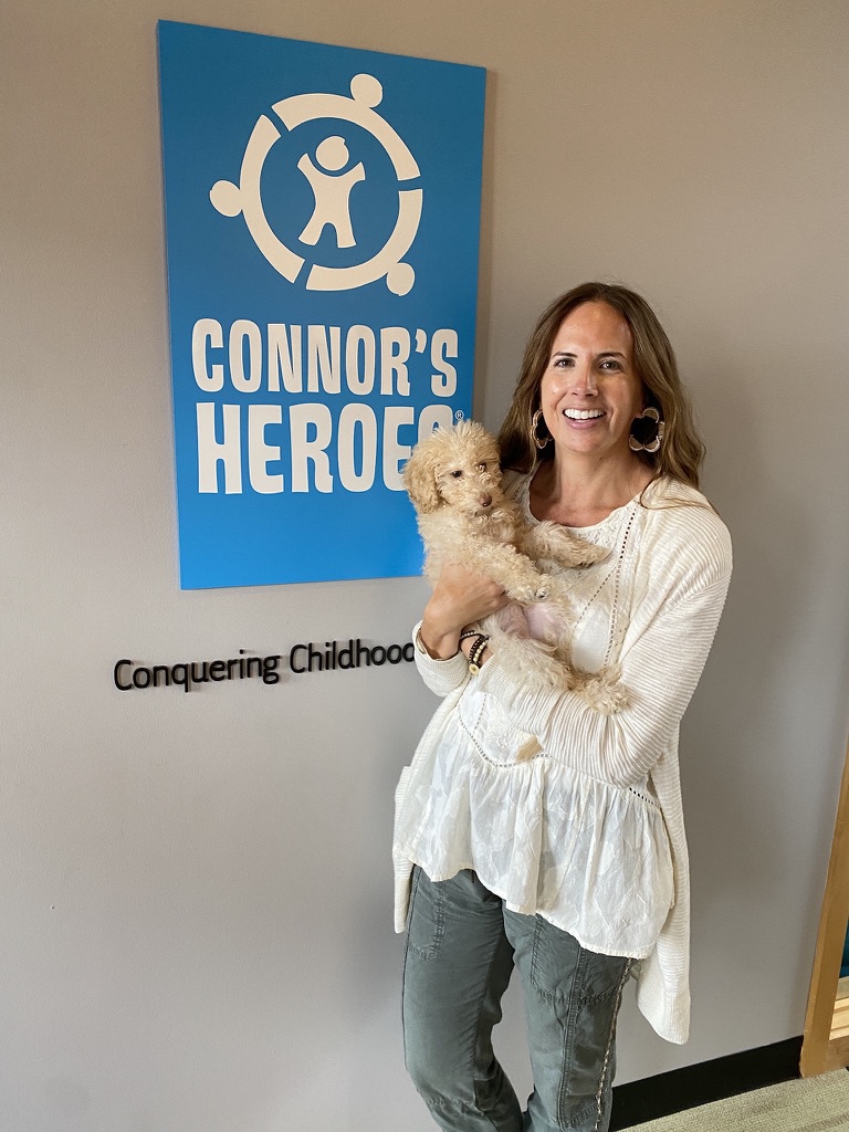 Woman holding a blonde puppy standing in an office in front of a sign for connors heroes