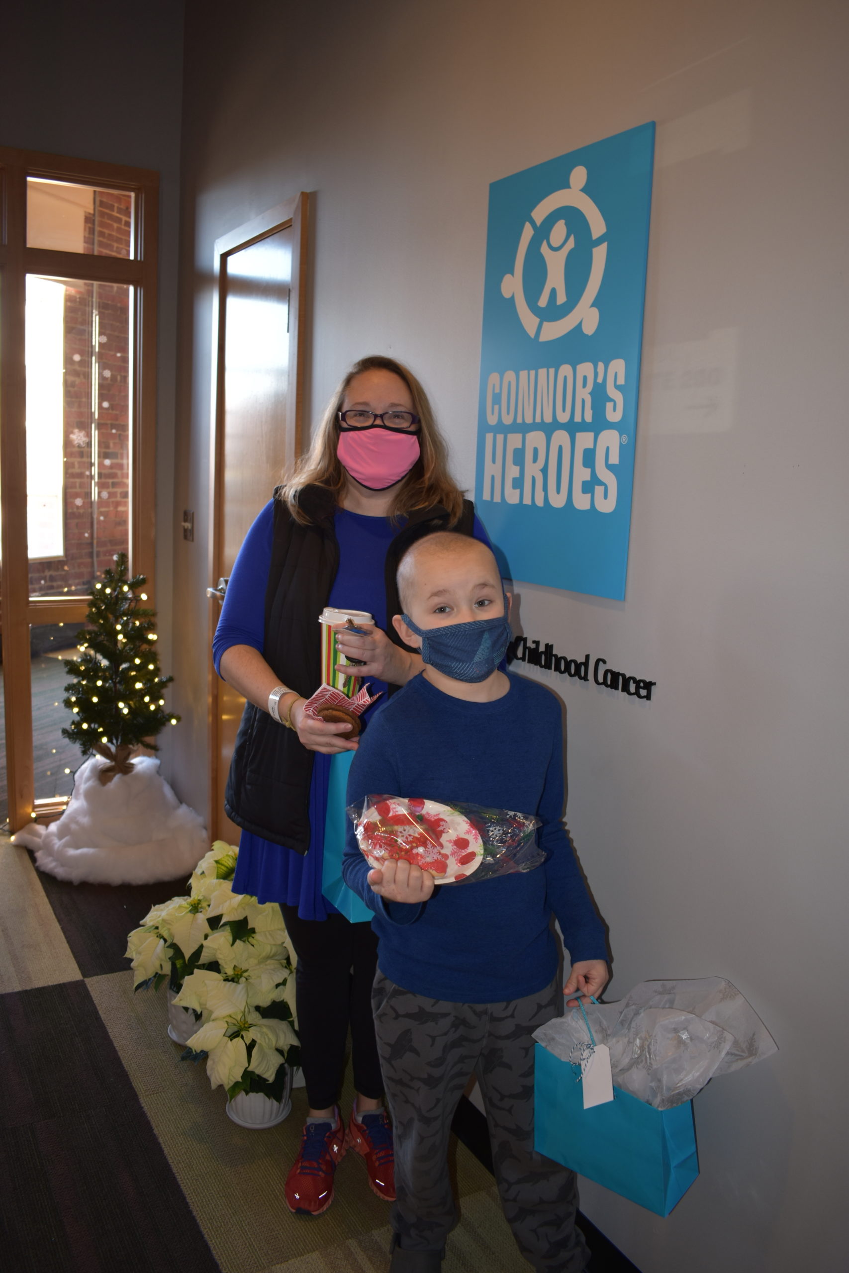 A mom stands with her son who is holding a plate of cookies and a gift bag full of presents