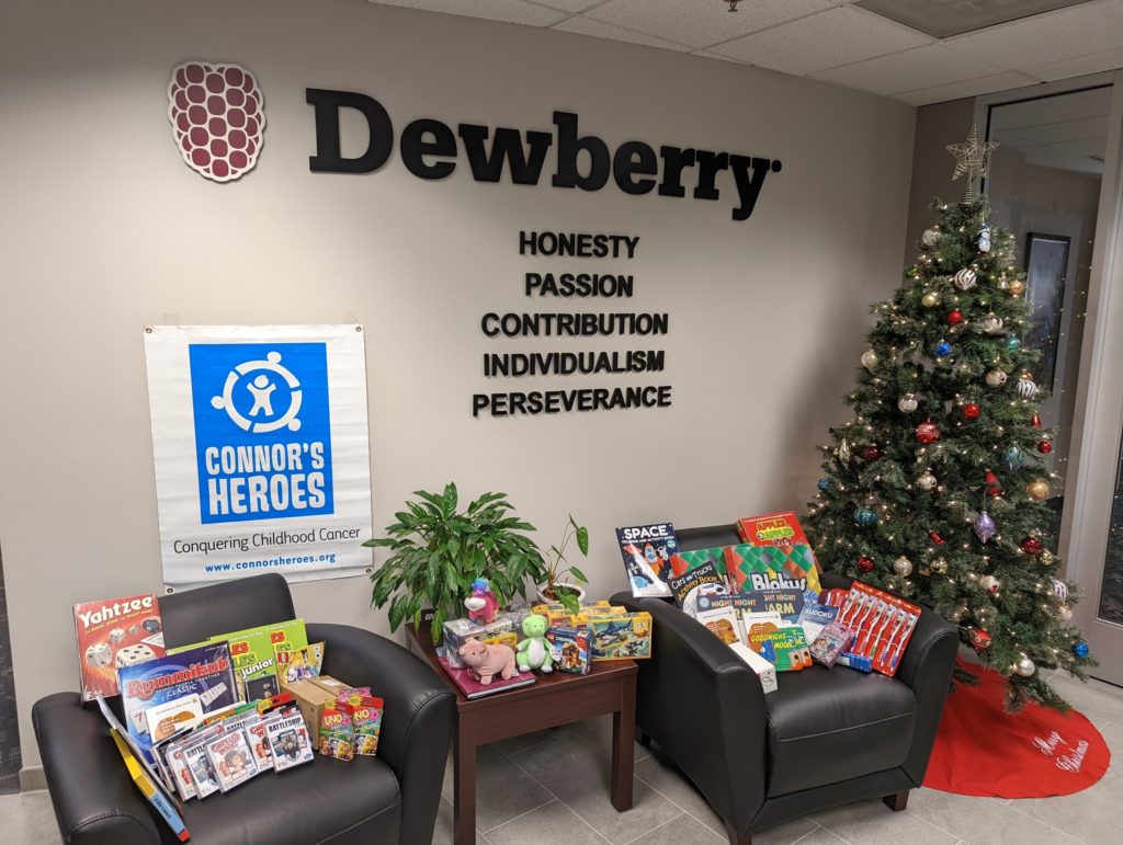Inside an office with the name Dewberry on the wall. There is a banner for Connors Heroes. There are two chairs and a table. Each has a pile of games and toys. They are next to a Christmas tree.