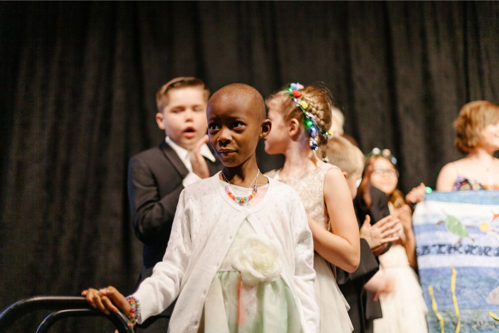 A child is standing on a stage behind her are a group of other children