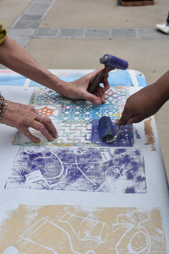 Three hands are on a large piece of cloth One hand is rolling purple ink while the other two hands hold a stencil