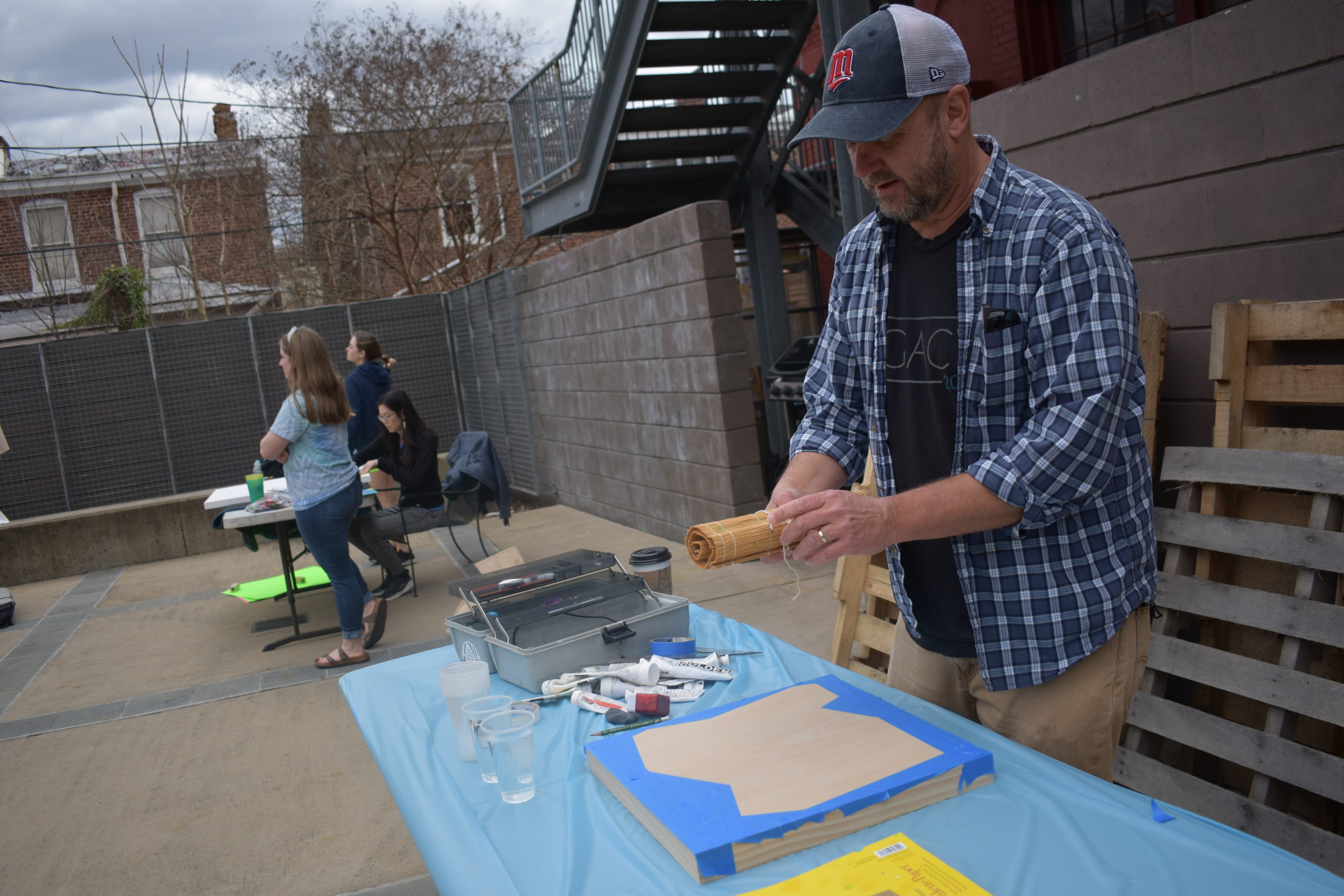 A man wearing a baseball cap is standing outdoors at a table with a wooden canvas and tubes of paint