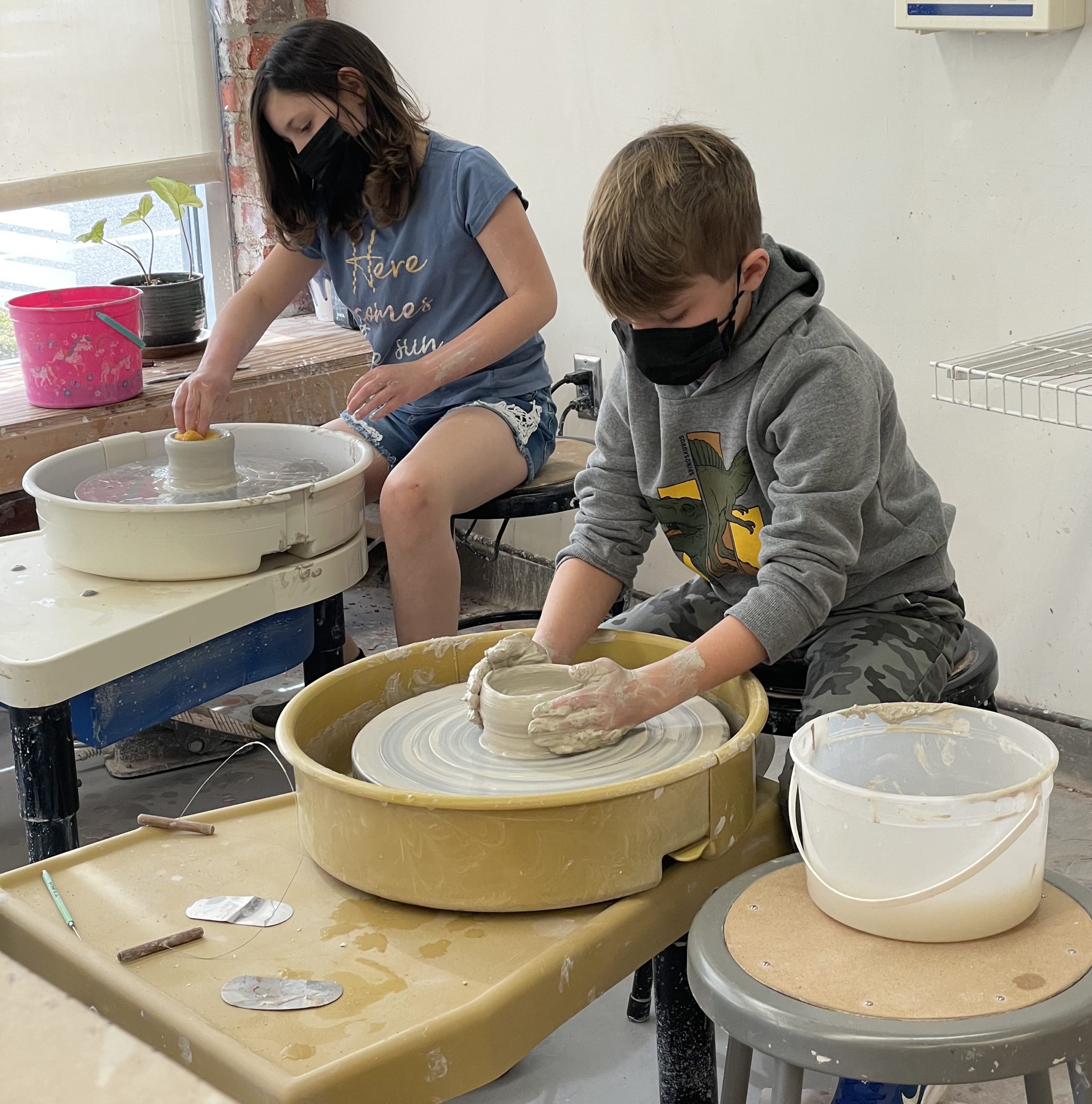 Two children sitting with pottery wheels shaping a piece of cl