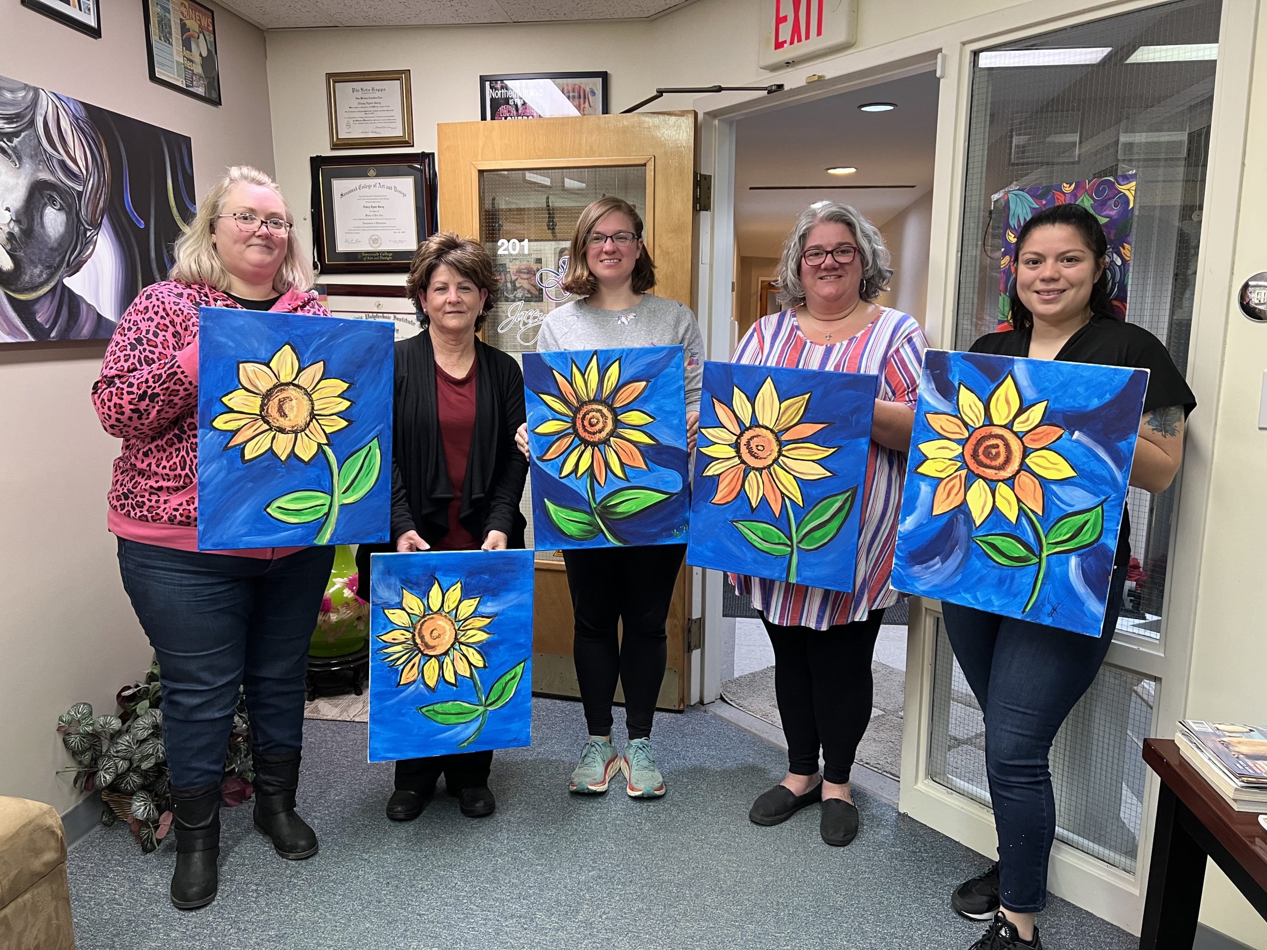 Five women standing together each one is holding a painting they did of a sunflower
