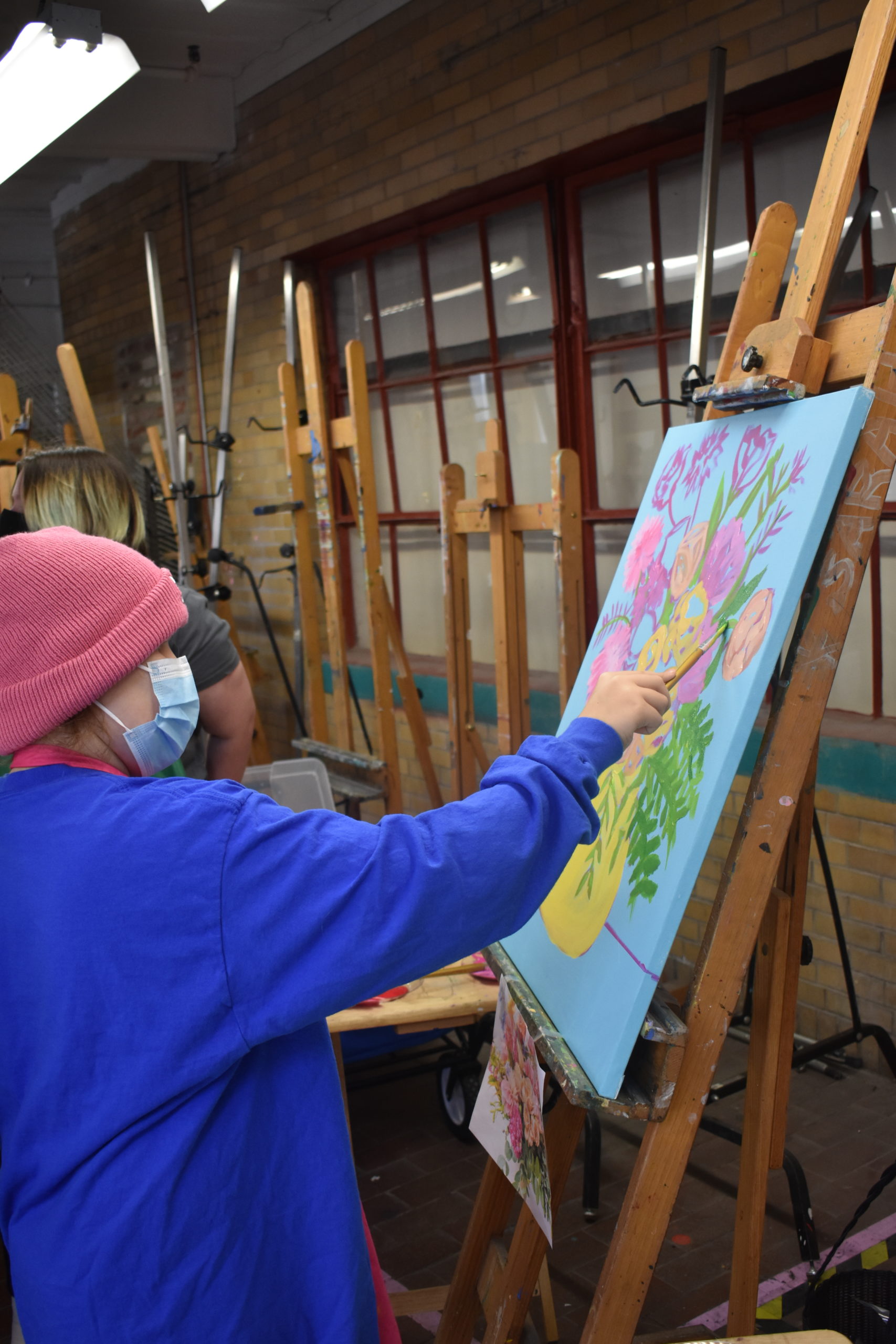 A child wearing a pink hat is painting a picture of a vase with flowers