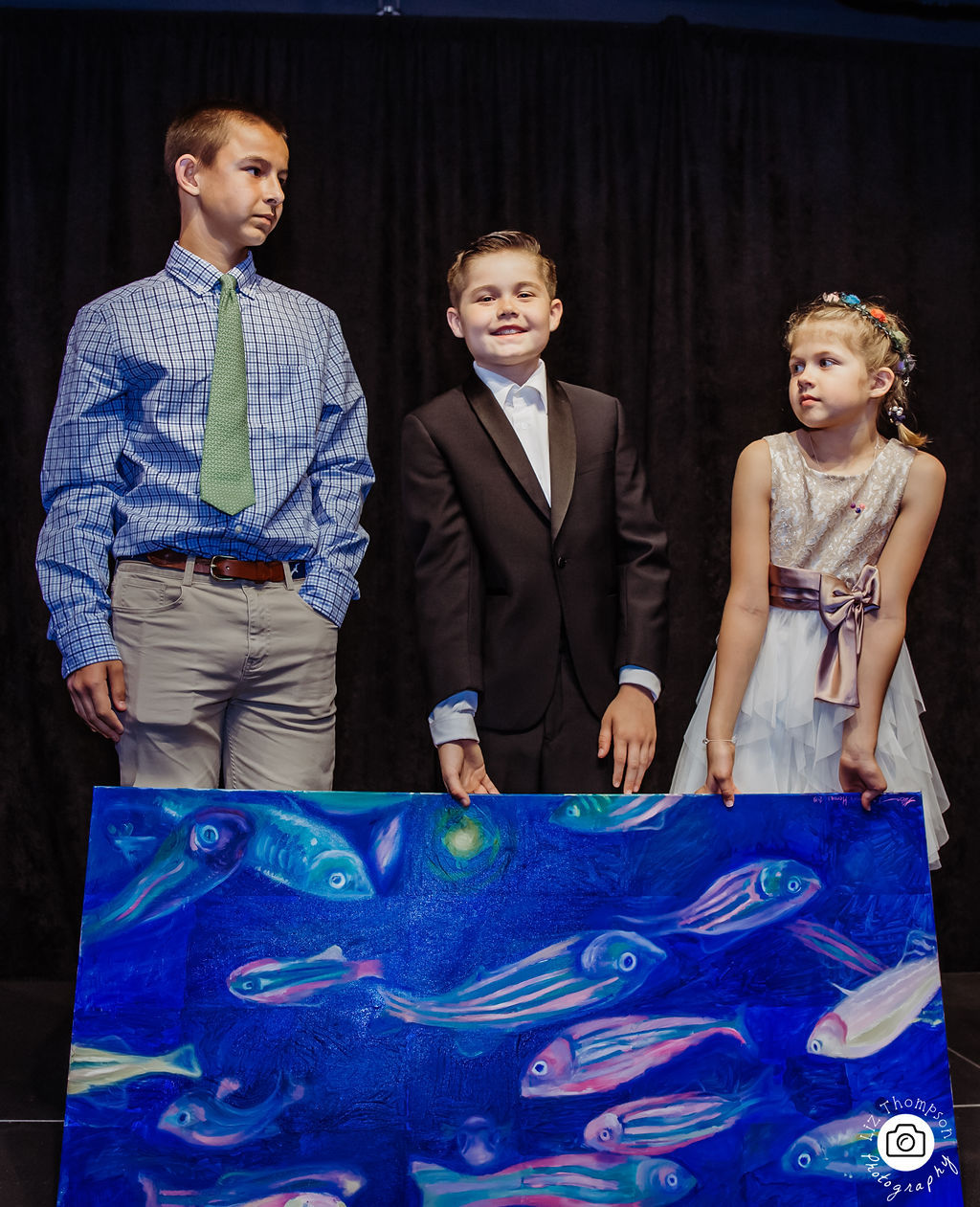 Three children standing on a stage holding a large painting of fish
