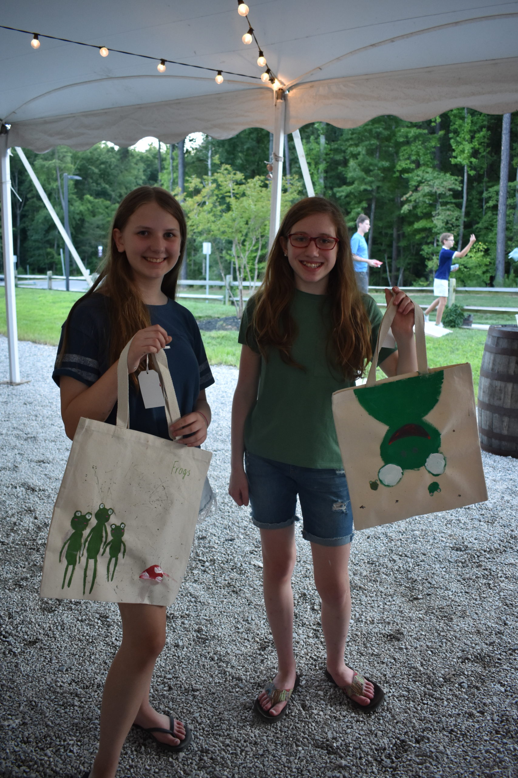 Two girls hold up tote bags they painted with frogs