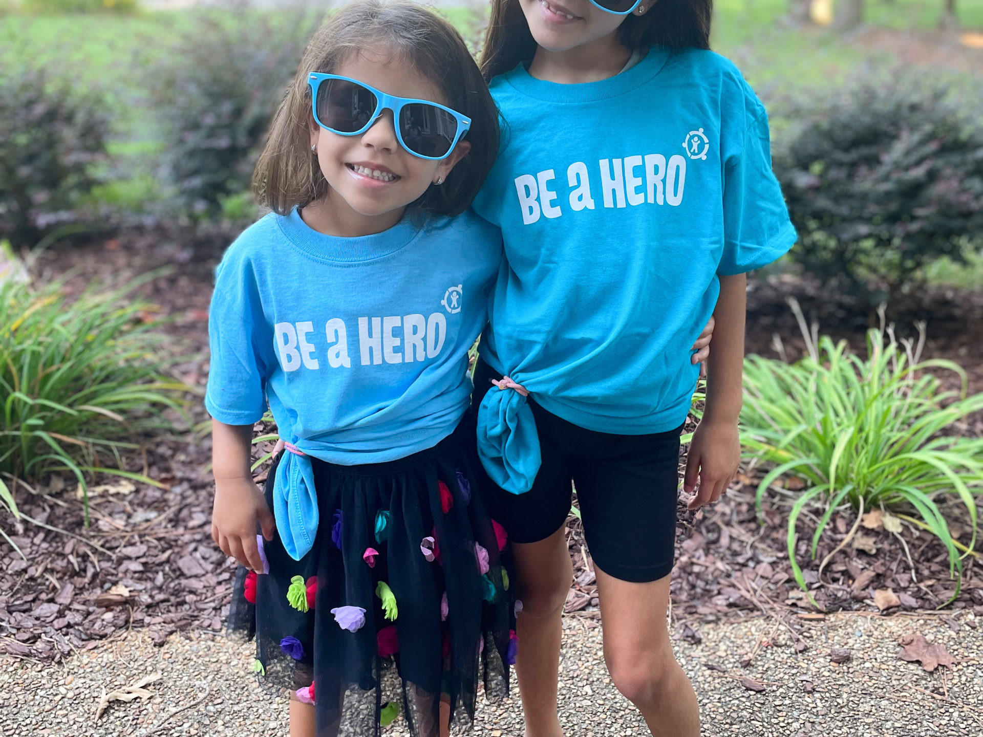 Two children standing together wearing bright blue shirts reading Be A Hero