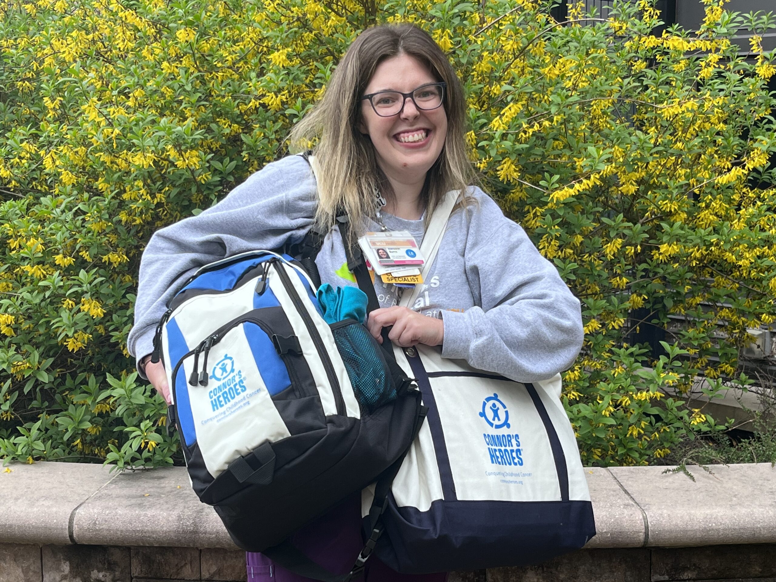 Meredith is standing outside in the hospital courtyard holding up a Heroes Bag and Backpack