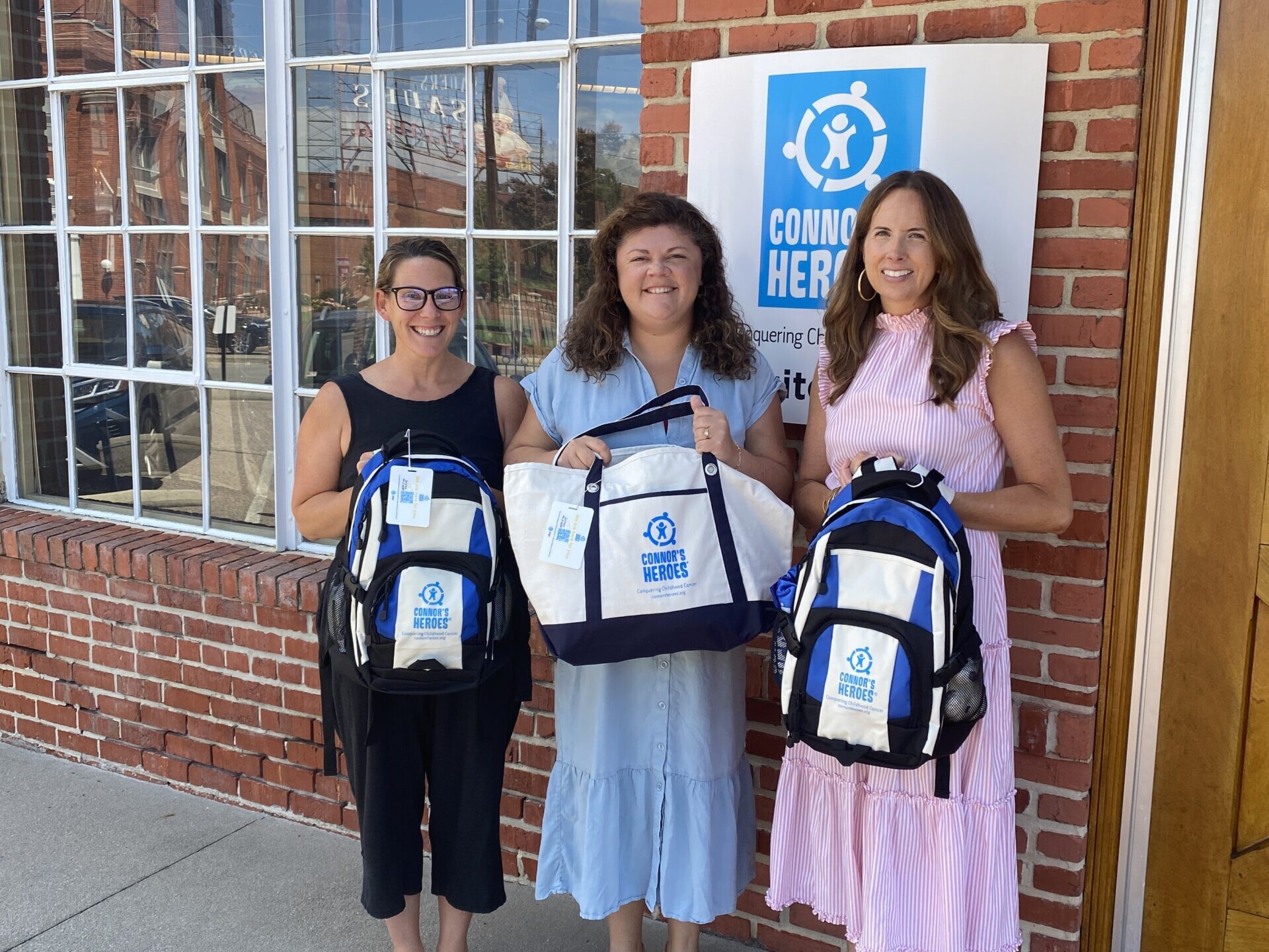 Connor's Heroes staff are holding up the heroes backpack and totebag
