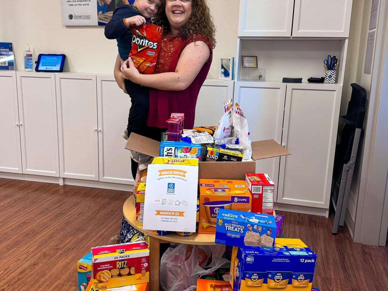 Celia holds her son Hal and they are standing in front a pile of snack boxes