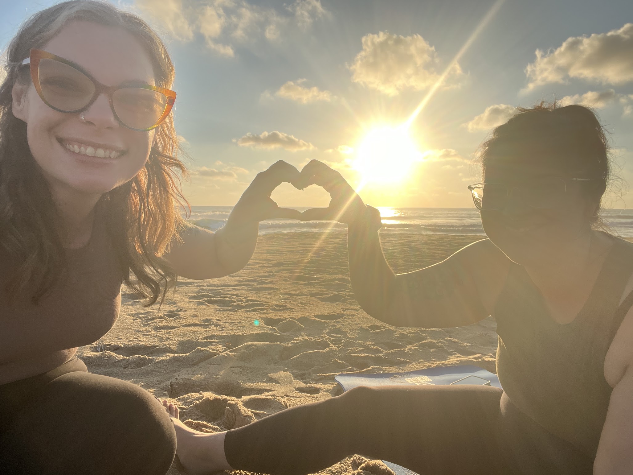 Two women make a heart with their hands while sitting on the beach